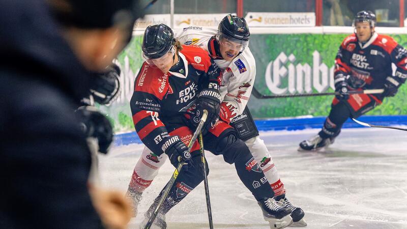 Zwei Eishockeyspieler in Aktion, einer in rotem Trikot mit schwarzer Hose, der andere in weißem Trikot mit roter Hose, kämpfen um den Puck auf dem Eis.