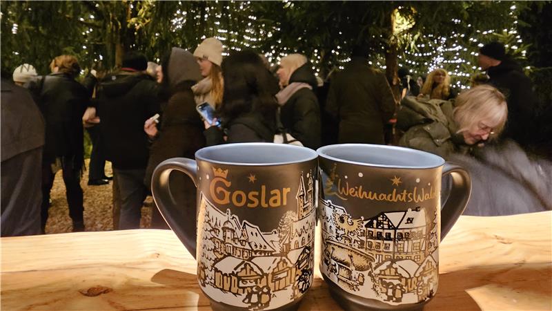 Zwei dunkelblaue Tassen mit goldener Aufschrift 'Goslar' und 'WeihnachtsWald' vor einem Weihnachtsmarkt mit beleuchteten Tannenbäumen und Menschen.