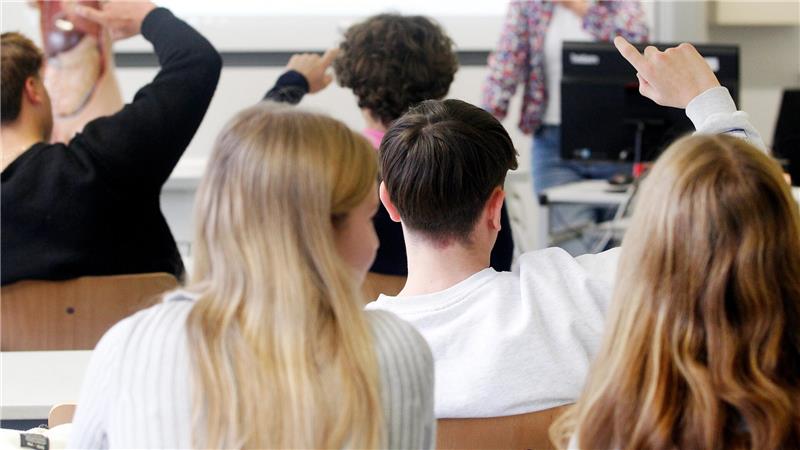 Mehrere Kinder sitzen in einem Klassenraum und heben die Hand, eine Frau steht vorne am Whiteboard und spricht