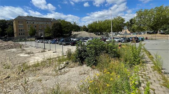 Blick auf eine Baustelle mit Schutt und Bauschutt im Vordergrund, dahinter ein Parkplatz mit mehreren Autos und im Hintergrund ein mehrstöckiges Gebäude sowie Bäume unter blauem Himmel