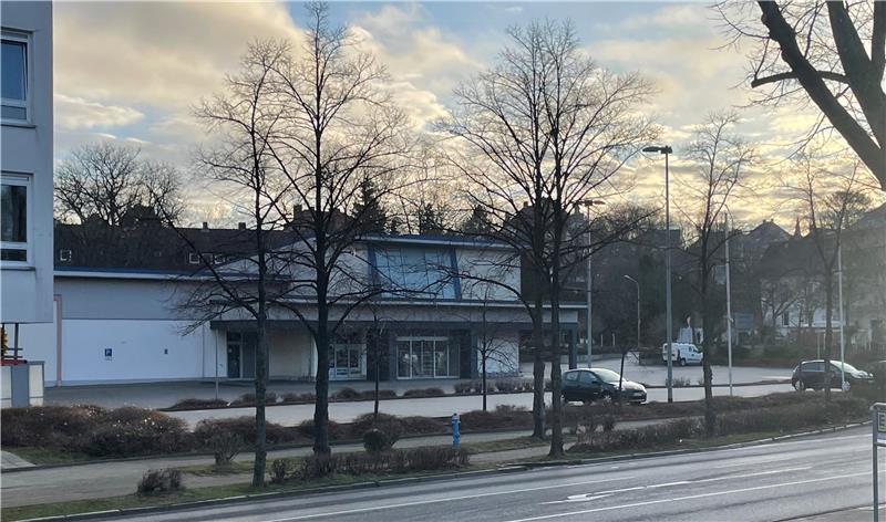 Straße mit Straßenbahnschienen, Bäumen ohne Blätter und einem weißen Gebäude im Hintergrund bei bewölktem Himmel