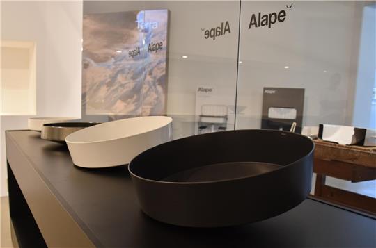 Drei unterschiedlich große, runde Waschbecken in Schwarz, Weiß und Grau auf schwarzer Theke in modernem Ausstellungsraum.