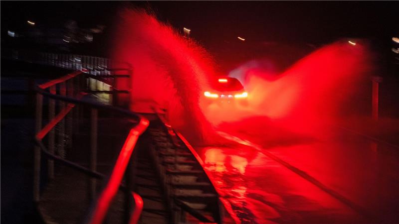 Wasser spritzt, während ein Auto über eine überschwemmte Straße zwischen Gleusdorf (Landkreis Haßberge, Unterfranken) und Busendorf (Landkreis Bamberg, Oberfranken) fährt.