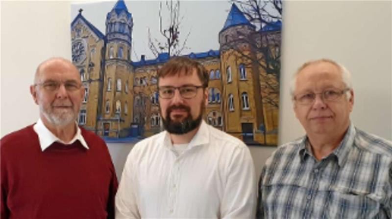 Drei Männer stehen vor einer Wand mit einem großen Bild eines historischen Gebäudes, sie tragen jeweils ein rotes, weißes und kariertes Hemd.