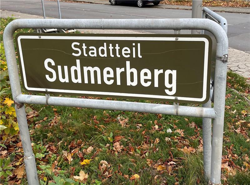 raunes Ortsschild mit weißer Schrift 'Stadtteil Sudmerberg' an einer Straße mit herbstlicher Vegetation im Hintergrund
