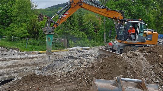 Bagger mit Hydraulikhämmer auf einer Baustelle vor bewaldetem Hügel