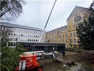 Ein Kran steht hinter einem schwarzen Container neben einem historischen gelben Backsteingebäude mit vielen Fenstern und einem großen Baum. Rotes Hubarbeitsgerät steht vor dem Container.