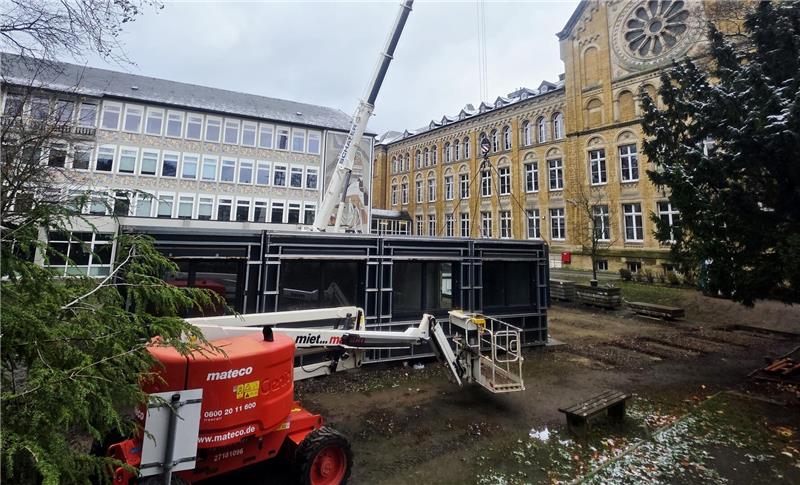 Ein Kran steht hinter einem schwarzen Container neben einem historischen gelben Backsteingebäude mit vielen Fenstern und einem großen Baum. Rotes Hubarbeitsgerät steht vor dem Container.
