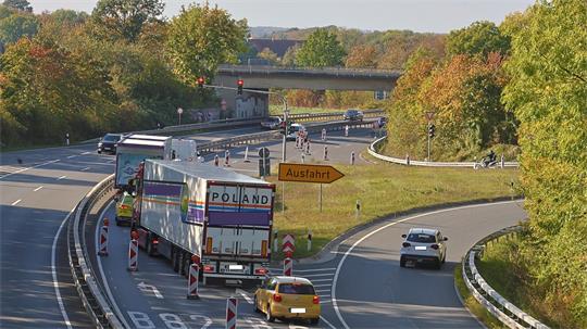 Mehrspurige Bundesstraße mit Baustellenabsperrungen und mehreren Fahrzeugen, darunter zwei Lastwagen. Eine gelbe Ausfahrtstafel weist auf die Ausfahrt hin; im Hintergrund führt eine Brücke über die Fahrbahn.