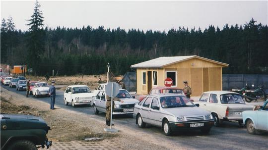 Das Bild zeigt eine Autoschlange auf einer Straße, eine Baracke und einen Mann, der die Autofahrer kontrolliert.