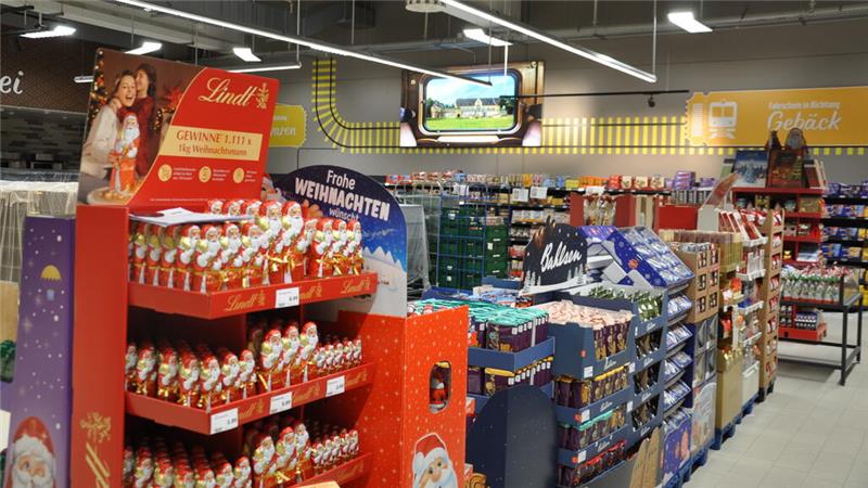 Supermarktregale mit weihnachtlicher Schokoladenwerbung und verschiedenen Süßwaren in Verpackungen, darüber ein Kaiserpfalz-Motiv und gelbe Beschilderung.