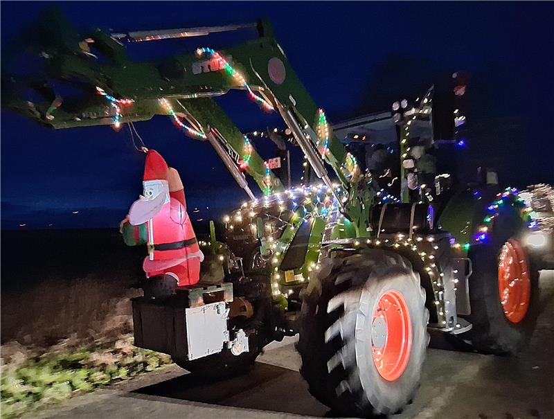 Traktor mit Frontlader, der mit Lichterketten geschmückt ist, trägt eine aufblasbare Weihnachtsmannfigur.