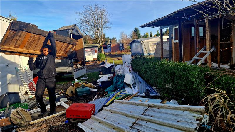 Das Bild zeigt eine Hütte, die gerade abgerissen wird. Ein junger Mann trägt Holz weg, rundherum liegt viel Schutt.