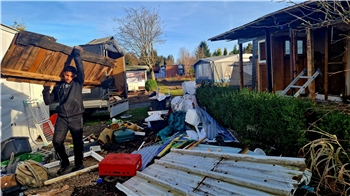 Das Bild zeigt eine Hütte, die gerade abgerissen wird. Ein junger Mann trägt Holz weg, rundherum liegt viel Schutt.