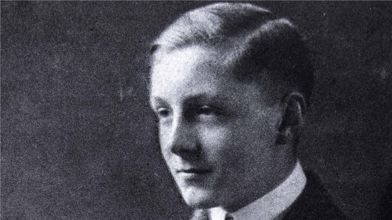 Junger Mann in Anzug mit verschränkten Armen sitzt vor dunklem Hintergrund