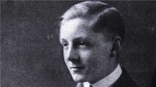 Junger Mann in Anzug mit verschränkten Armen sitzt vor dunklem Hintergrund