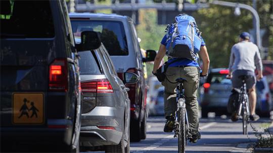 Zwei Radfahrer fahren auf einer Straße zwischen parkenden und fahrenden Autos