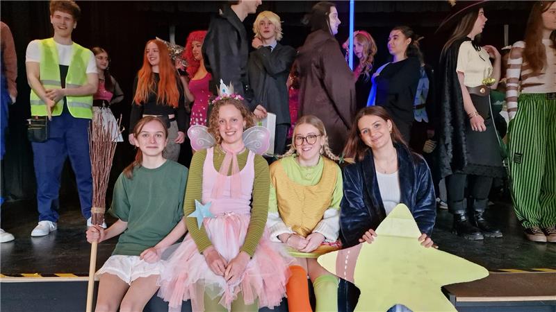 Gruppe von Menschen in bunten Kostümen auf einer Bühne, einige sitzen vorne mit Requisiten wie einem Stern und einem Stab, andere stehen im Hintergrund.