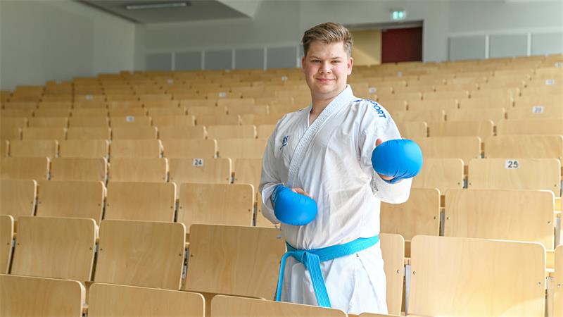 Junger Mann im Karateanzug mit blauen Handschuhen steht in einem leeren Hörsaal und lächelt in die Kamera.
