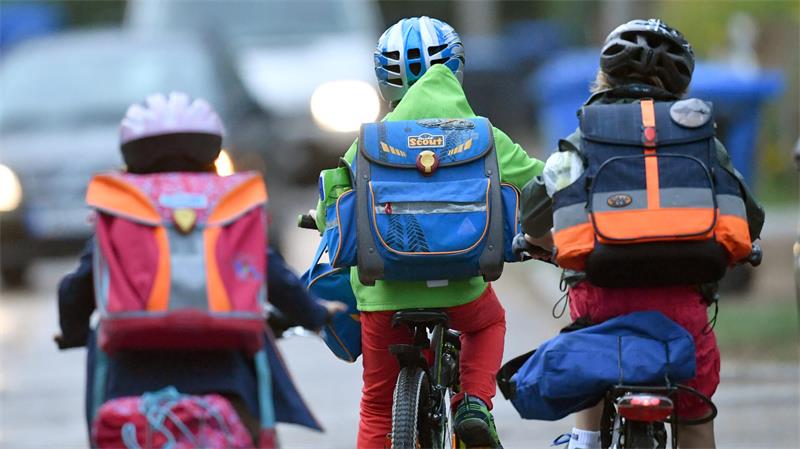 Drei Kinder mit Schulranzen und Fahrradhelmen fahren auf Fahrrädern auf einer Straße