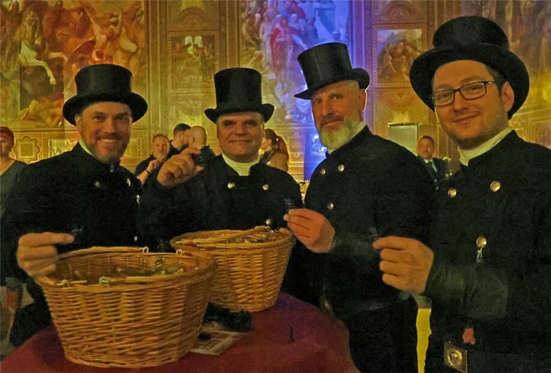 Vier Männer in schwarzer traditioneller Kleidung und Zylinderhüten stehen um einen Tisch mit zwei großen Weidenkörben in einem prunkvollen Saal mit Wandgemälden.