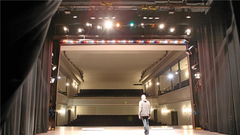 Person mit Kapuzenpullover steht auf einer Bühne und blickt in einen leeren Zuschauerraum eines Theaters