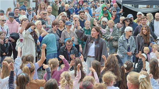 Menschenmenge bei einer Veranstaltung mit vielen Kindern im Vordergrund, die die Arme heben, und Erwachsenen im Hintergrund vor Marktständen