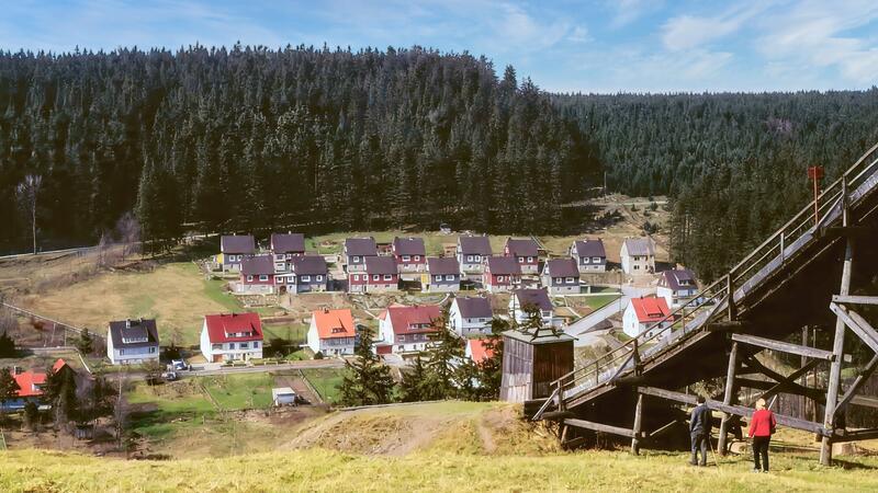 Blick auf die Altenauer Bornkappe mit Reihenhäusern vor einem dichten Wald und einer Holzskisprungschanze, an der zwei Personen stehen.