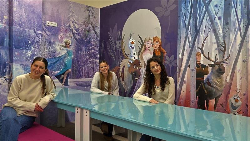 Drei Personen sitzen an einem langen, türkisfarbenen Tisch in einem Raum mit Wandbildern der Figuren aus dem Film "Die Eiskönigin".