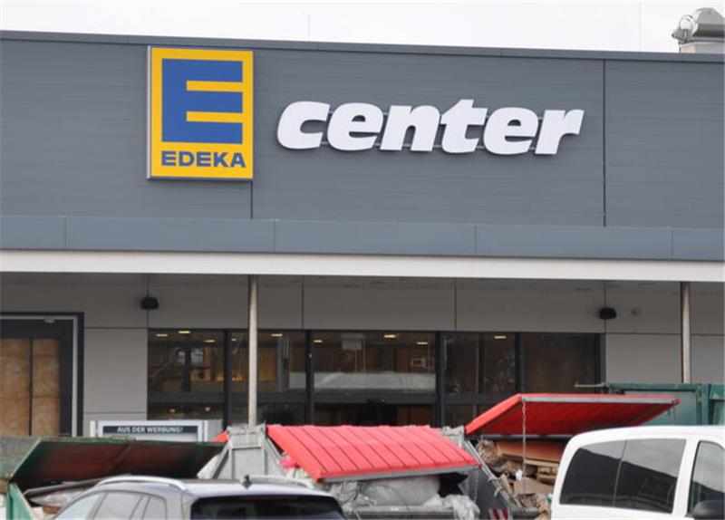 Fassade eines Edeka-Centers mit grauer Außenwand und gelb-blauem Logo, davor stehen mehrere geparkte Fahrzeuge und Container.