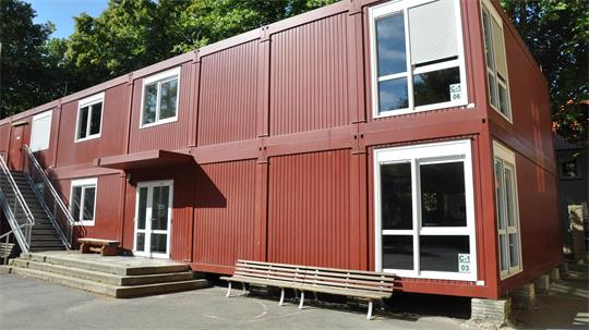 Zweistöckiges Gebäude aus roten Containermodulen mit weißen Fenstern und Türen, davor zwei Holzbänke und eine Außentreppe links
