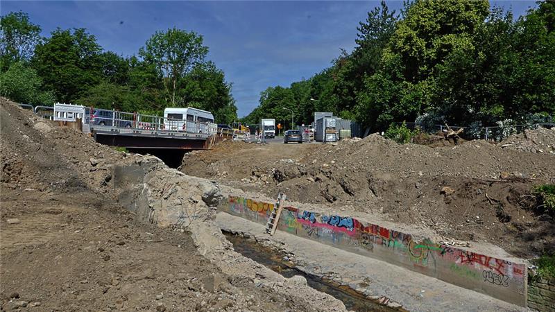 Baustelle mit ausgehobener Erde und einem betonierten Kanal mit Graffiti, im Hintergrund Bäume und Fahrzeuge auf einer Straße
