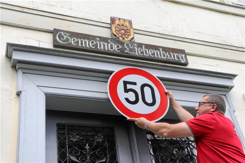 Wie es sich für einen 50. Geburtstag gehört – das obligatorische Verkehrsschild: Bürgermeister Alf Hesse klettert auf die Leiter und hält für das Foto ein Tempo-50-Zeichen an den Eingang der Gemeindeverwaltung. Das alte Holzschild ist übrigens deutlich sichtbar in die Jahre gekommen – es könnte dort seit den 1970er Jahren, als die Verwaltung in das Haus einzog, so unverändert hängen, meint der Bürgermeister. Foto: Gereke