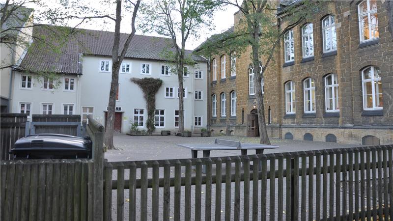 Pausenhof, der von zwei Gebäuden ungeben ist, einem weißen mit Satteldach und einem braunen mit hohen Fenstern, davor ein Holzzaun und mehrere Bäume.