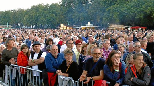 Große Menschenmenge vor Absperrung im Freien, im Hintergrund Bäume und Verkaufsstände.