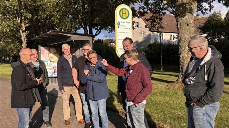 Gruppe von acht Personen steht an einer Bushaltestelle auf einem Gehweg neben einer Straße mit Bäumen und Häusern im Hintergrund