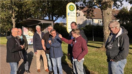 Gruppe von acht Personen steht an einer Bushaltestelle auf einem Gehweg neben einer Straße mit Bäumen und Häusern im Hintergrund