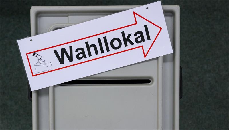 Eine Wahlurne. Darauf liegt ein Zettel mit einem Pfeil nach rechts und der Aufschrift "Wahllokal". 