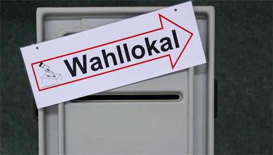 Eine Wahlurne. Darauf liegt ein Zettel mit einem Pfeil nach rechts und der Aufschrift "Wahllokal". 