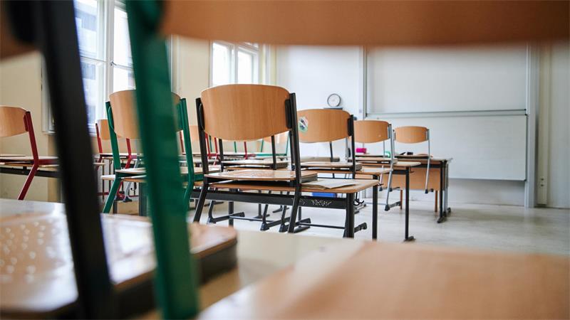 Leerer Klassenraum mit mehreren Holzstühlen und Tischen vor großen Fenstern und einer weißen Tafel.
