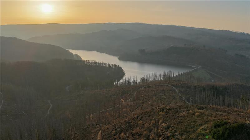 Landschaft mit Fluss, der sich durch hügeliges Gelände schlängelt, im Hintergrund Sonnenuntergang.