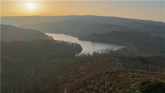 Landschaft mit Fluss, der sich durch hügeliges Gelände schlängelt, im Hintergrund Sonnenuntergang.