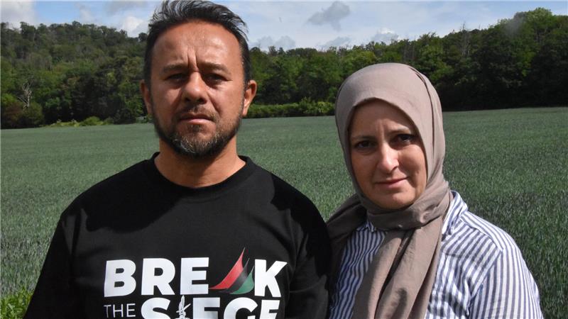 Zwei Personen stehen nebeneinander auf einem Feld vor bewaldetem Hintergrund, eine trägt ein schwarzes T-Shirt mit weißer und roter Aufschrift, die andere einen gestreiften Oberteil und ein helles Kopftuch.