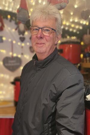 Glühwein & Co. auf dem Weihnachtsmarkt: Was kostet was? Willi Robben steht seitlich zur Kamera in einer schwarzen Jacke während im Hintergrund leuchtende Girlanden und Weihnachtsschmuck zu sehen ist.