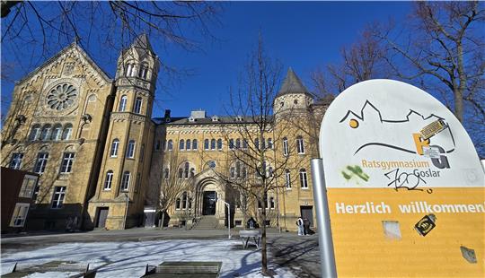 Historisches Schulgebäude mit Türmen und Rosettenfenster im Hintergrund sowie einem Schild mit der Aufschrift 'Ratsgymnasium Goslar Herzlich willkommen' im Vordergrund