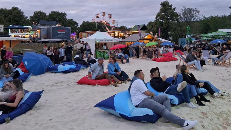 Menschen sitzen auf bunten Sitzsäcken im Sand auf einem Festivalgelände mit Fahrgeschäften und Ständen im Hintergrund