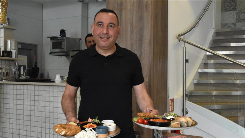 Mann in schwarzem T-Shirt trägt Tabletts mit verschiedenen Speisen in einem Restaurant mit Treppe im Hintergrund.