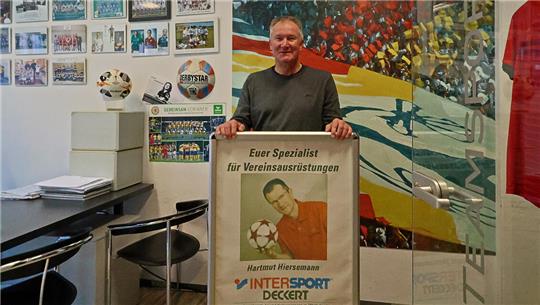 Person steht in einem Büro und hält ein Plakat mit der Aufschrift 'Euer Spezialist für Vereinsaustüstungen Hartmut Hiersemann Intersport Deckert'. Im Hintergrund bunte Wandgestaltung und mehrere gerahmte Fotos an der Wand.