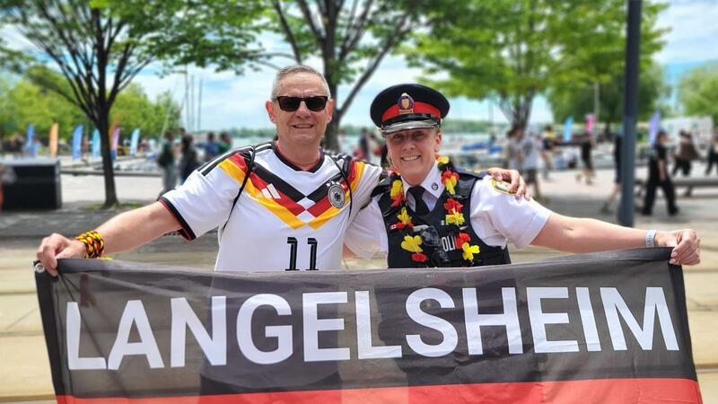 Zwei Personen in Deutschland-Trikots halten ein Banner mit der Aufschrift 'LANGELSHEIM' vor einer Baum- und Menschenkulisse im Freien