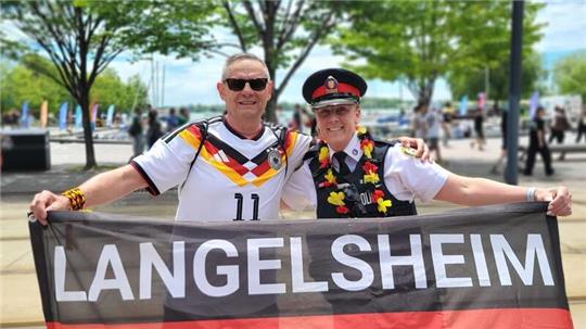 Zwei Personen in Deutschland-Trikots halten ein Banner mit der Aufschrift 'LANGELSHEIM' vor einer Baum- und Menschenkulisse im Freien